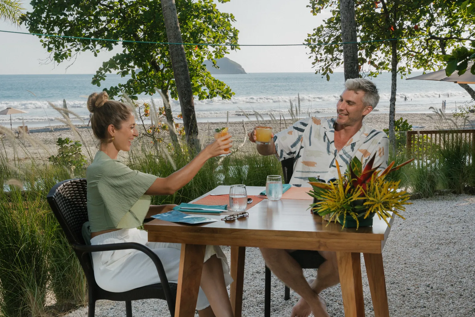 A couple toasting with cocktails at a table facing the beach